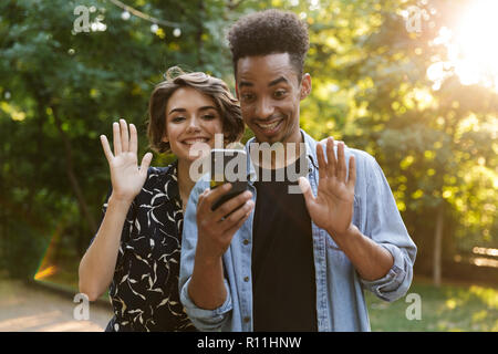 Happy multiethnic couple at the park, waving hands to mobile phone