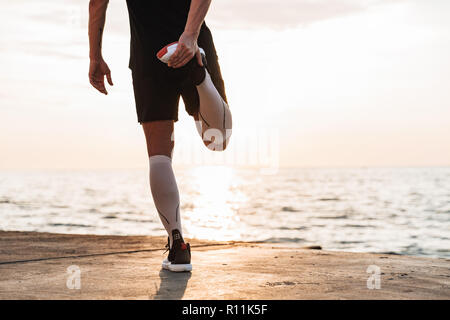 Cropped photo of handsome strong young sportsman outdoors at the beach make stretching exercises. Stock Photo