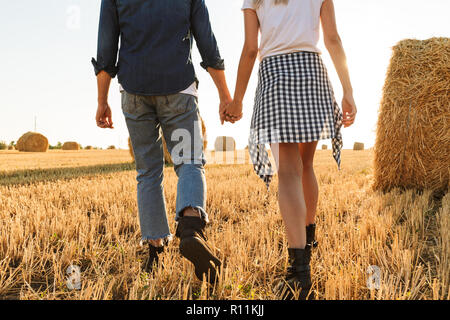 Cropped photo of young guy and girl walking through golden field with bunch of haystacks during sunny day Stock Photo