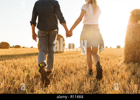 Cropped photo of guy and girl walking hand by hand through golden field with bunch of haystacks during sunny day Stock Photo