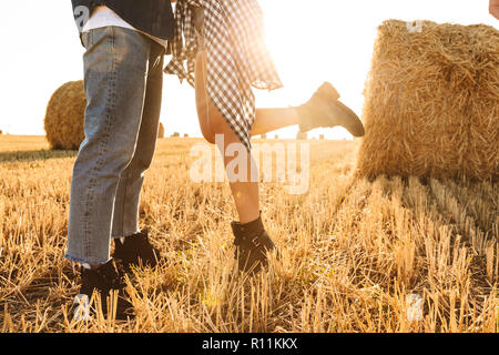 Cropped photo of guy and girl walking through golden field with bunch of haystacks and kissing during sunny day Stock Photo