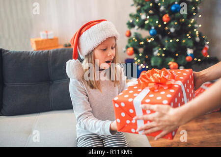 Amazed child holds present and looks at it. She keeps mouth opened. Girl wears Christmas hat. Adult supports her by holding present with hands. They a Stock Photo