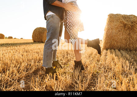 Cropped photo of guy and girl walking through golden field with bunch of haystacks and hugging during sunny day Stock Photo