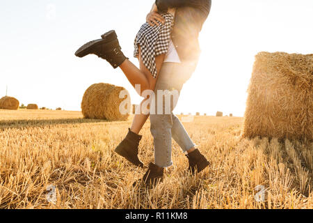 Cropped photo of guy and girl in love walking through golden field with bunch of haystacks and hugging during sunny day Stock Photo