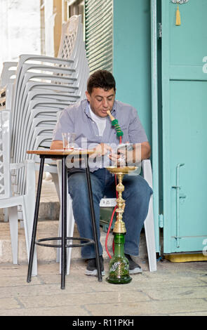 An Arab man simultaneously smokes a hookah pipe and texts on his cell phone in the Muslim Quarter of the old City of Jerusalem in Israel. Stock Photo