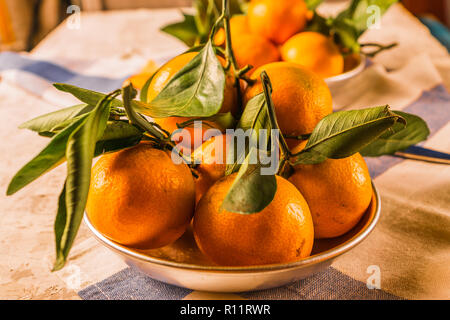 Flat lay view of Orange ripe mandarins fruit with green leaves, in a bowl, on a blue and white napkin, over rustic white wooden table. Stock Photo