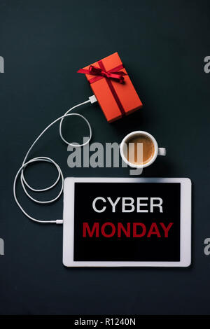 high angle view of a tablet with the text Cyber Monday in its screen, connected to a gift by a cable, and a cup of coffee, placed on a dark surface Stock Photo