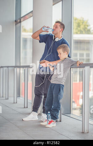 Little boy and his father taking a break, drinking water and listening to the music together in a sports center. Father and son spend time together an Stock Photo