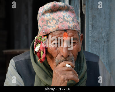 Elderly Nepali Newari man with a red tilaka mark on his forehead wears the traditional Nepali dhaka topi hat and smokes a cigarette. Stock Photo