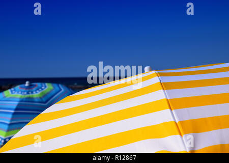 different colorful striped umbrellas on beach, yellow and white striped parasol in front of azure sky as a symbol of vacation Stock Photo