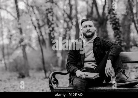 A handsome young hipster man sitting on a bench with a book and daydreaming. Stock Photo