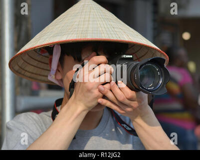 Young Vietnamese street photographer wears an Asian conical hat and takes a photo with his Canon DSLR camera. Stock Photo