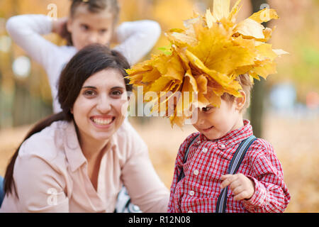 Mother puts yellow fallen leaves on son head. Happy family is in autumn city park. Children and parents. They posing, smiling, playing and having fun. Stock Photo