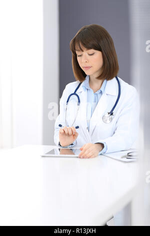 Female doctor typing on laptop computer while sitting at the white table in hospital office. Physician at work. Medicine and healthcare concept Stock Photo