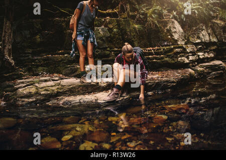 Two women hikers by a water pond in mountain. Woman sitting on rock and putting her hand in water with friend standing by and looking. Stock Photo