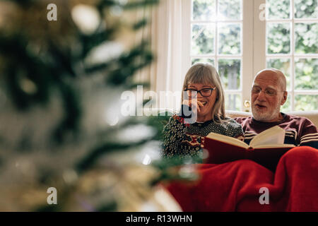 Old woman laughing while her husband reads a book. Senior couple reading a book sitting on couch at home with a christmas tree in the foreground. Stock Photo