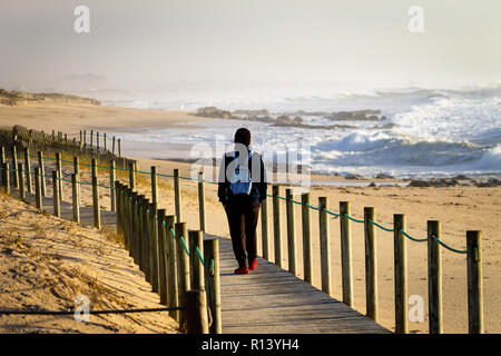 Young man walks on boardwalk near the sand and the ocean. Backpack. Back View. Clear Day. Stock Photo