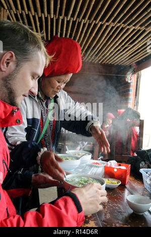 Red dzao woman serves pho ga (chicken noodle soup) to a German tourist at her village eatery near Sapa, Vietnam Stock Photo