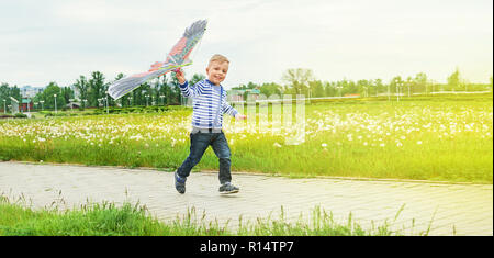 Little boy running across park with toy kite flying. Caucasian child playing on spring day. Lifestyle kid actively recreation .Copy space for text,blank background. Summer,outdoors, green grass. Stock Photo