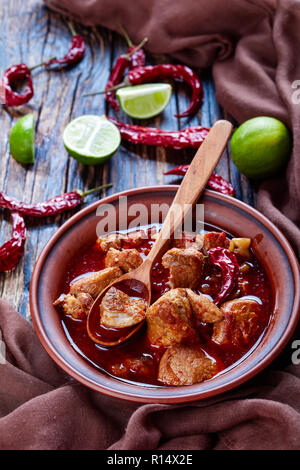 Carne Adobada or hearty Red Chile and Pork Stew in a clay bowl with spoon on an old wooden table with lime slices and dried chile, mexican cuisine, ve Stock Photo
