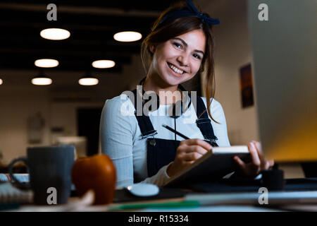 Image of happy smiling young woman designer sitting in office working reading book taking notes at night. Stock Photo