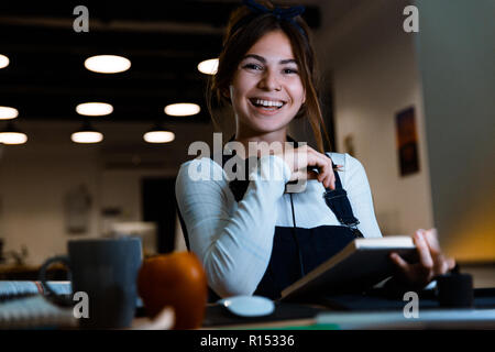 Image of happy smiling young woman designer sitting in office working reading book taking notes at night. Stock Photo