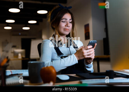 Image of confused young woman designer sitting in office working using mobile phone at night. Stock Photo