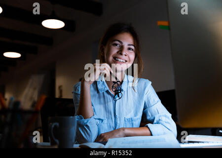 Image of happy cheerful young woman graphic designer using pc computer working at night in office looking camera. Stock Photo