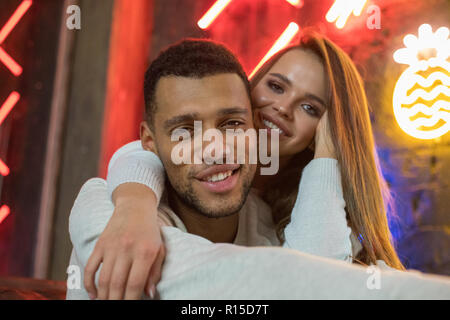 Happy multiracial couple sits embracing on the background of neon lamps. Stock Photo