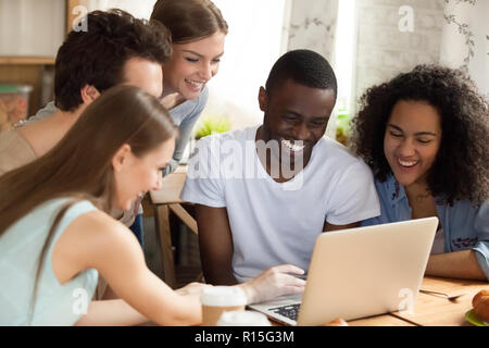 Black african guy with diverse friends watching comedy movie funny videos online using computer sitting together at desk. Friendship between multiraci Stock Photo
