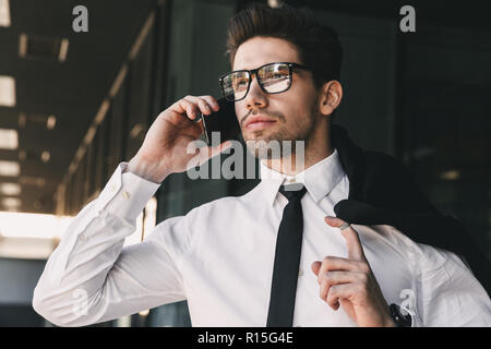 Portrait closeup of young businessman dressed in formal suit standing outside glass building with jacket over his shoulder and talking on mobile phone Stock Photo