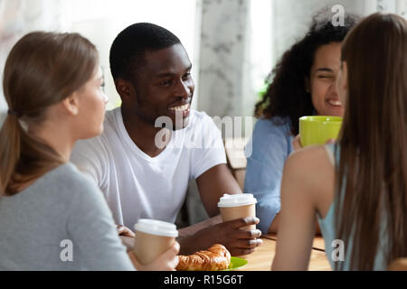 Smiling black man sitting at table surrounded by friends diverse girls sitting chatting together drinking coffee tea having pleasant conversation in c Stock Photo
