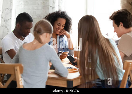 Positive diverse friends sitting around table in cafe. Multiracial students browsing internet communicating chatting online holding mobile phones. Wir Stock Photo