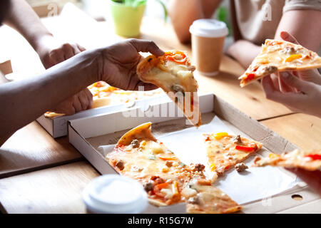 Diverse people having lunch eating fast junk food. Close up hands diverse multi-ethnic students holding pieces of pizza sitting at table in cafe. Unhe Stock Photo