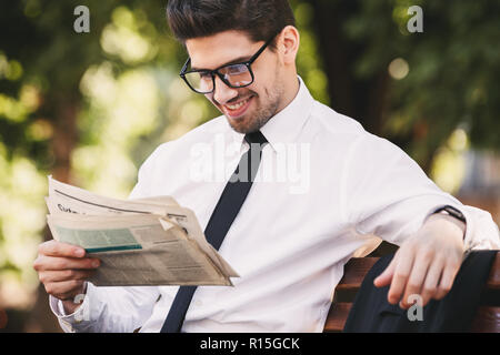 Photo of successful man in businesslike suit sitting on bench in green park and reading newspaper during sunny day Stock Photo