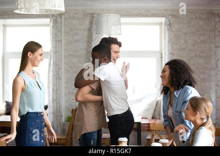 Diverse students gathered together during lunch. Guys buddies best friends hugging greeting each other at meeting in cafe surrounded by attractive mul Stock Photo