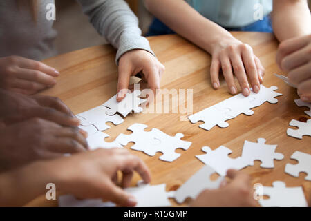 Close up diverse male female hands multiracial people assembling puzzle on wooden table cogitating making decision together. Team building and teamwor Stock Photo