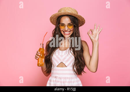 Photo of caucasian woman 20s wearing sunglasses and straw hat drinking lemonade from glass bottle isolated over pink background Stock Photo