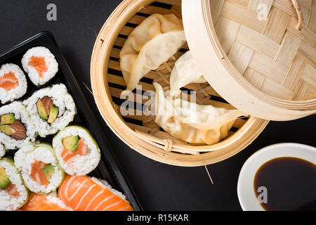 Dumplings or gyoza served in traditional steamer and sushi on black background. Flat lay top-down composition