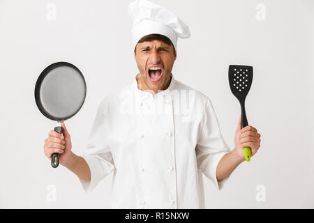 Image of screaming angry young chef man standing isolated over white wall background holding frying pan looking camera. Stock Photo