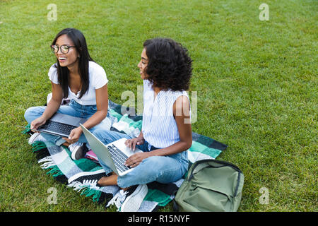 Two happy young girls students sitting on a grass at the campus, studying with laptop computers Stock Photo