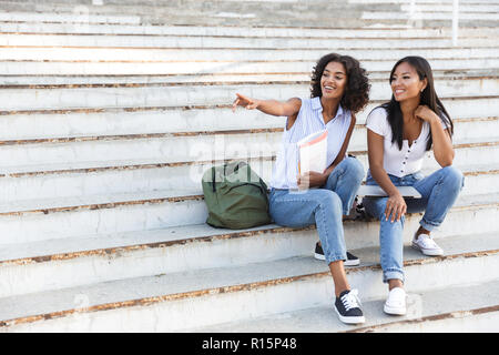 Two smiling young girls students sitting on steps outdoors, pointing away Stock Photo