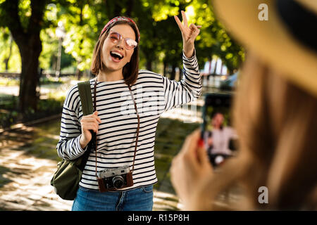 Photo of young happy ladies friends walking outdoors with backpacks take a photo by mobile phone. Stock Photo