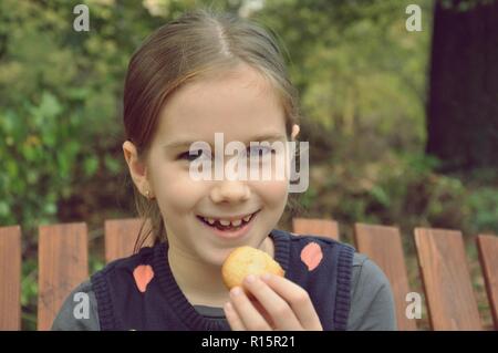 Caucasian 8 years old child, girl, sitting on a bench, smiling and eating a biscuit in the park Stock Photo