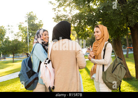 Group of three happy muslim women students carrying backpacks and books, standing outdoors, chatting Stock Photo