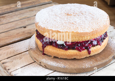 Victoria Sponge cake with fruit jam and vanilla cream on rustic wooden cake stand dusted with icing sugar. Stock Photo