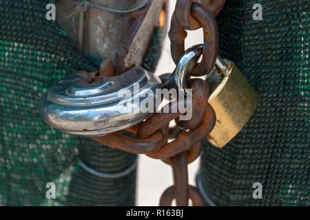 A close up view of two locks and a old rusted chain securing a matal gate with green mesh on Stock Photo