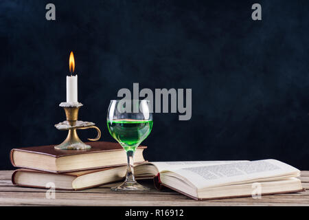 Absinthe drink in glass and books with candle in candlestick burning on retro wooden desk and smoky dark background. Green fairy inspiration and muse Stock Photo