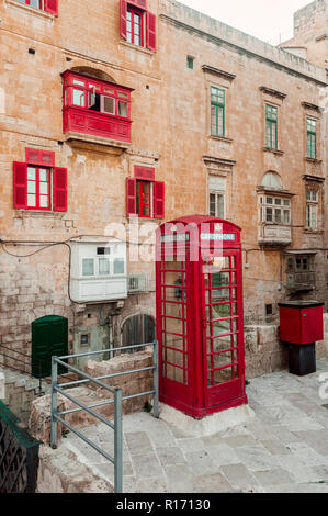 Old vintage maltese red telephone box with red traditional Maltese balcony and shuttered windows, against yellow limestone walls, Valletta, Malta Stock Photo