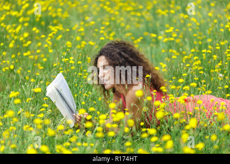 Cute young woman with curly hair lying reading a book in a wildflower meadow full of colorful yellow summer flowers as she relaxes in the tranquility  Stock Photo